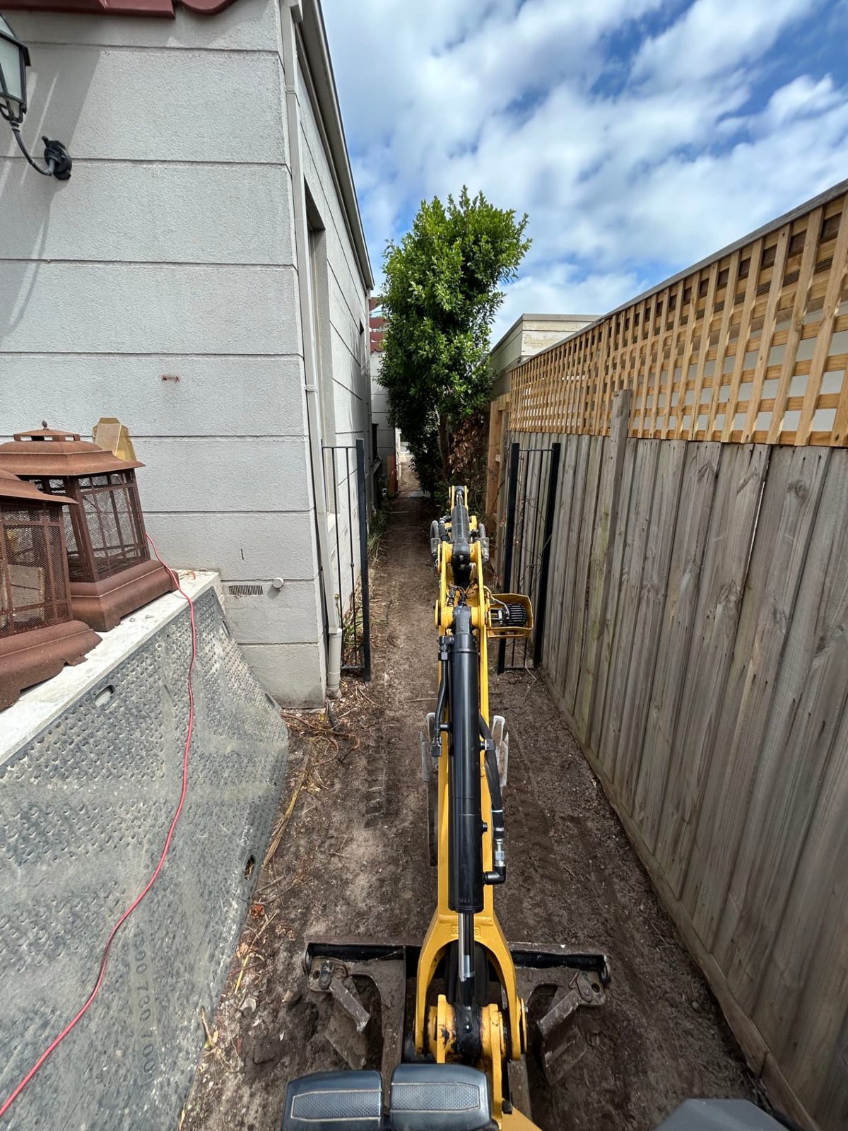Rigid Earthworks compact excavator working in a narrow side path between a house and boundary fence